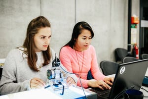 two young women using a computer to operate a robot. One in a pink jumper, the other in a yellow shirt